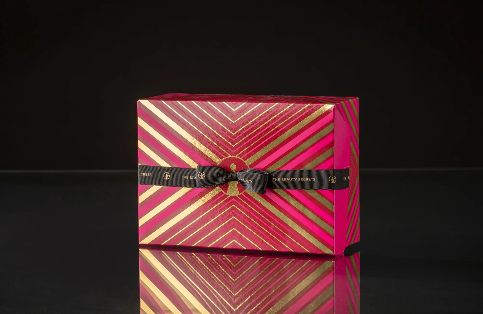 Pink and gold striped Beauty Secrets gift box finished with a printed ribbon and bow, on a black reflective ground